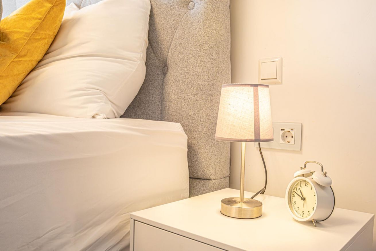 Ein ordentlich bezogenes Bett mit Kissen, einer Nachttischlampe und einem an der Wand befestigten Wecker auf einem weißen Nachttisch.