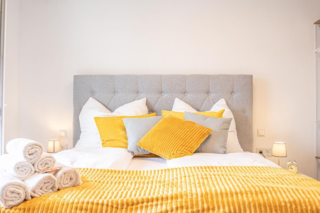 Een keurig opgemaakt bed met een grijs gestoffeerd hoofdbord, witte en gele kussens en een gele gestreepte deken.