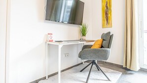 Ein modernes Zimmer mit flachbildfernseher an der Wand, einem Schreibtisch mit Computer, einem Stuhl, einer Topfpflanze und einem Gemälde.