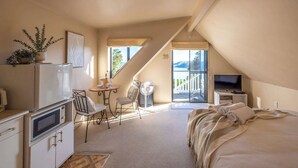 Une pièce compacte et bien éclairée, comprenant un lit, un bureau, une chaise, une petite table, un téléviseur et une fenêtre donnant sur l’extérieur.