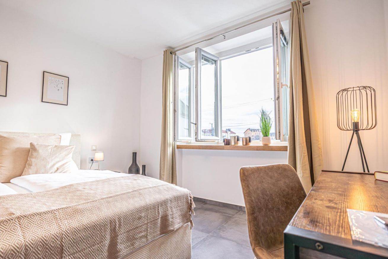 A hotel room with a bed, a desk, a chair, a window with curtains, and a lamp.