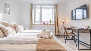 Habitación de hotel con cama, escritorio, silla, televisión, ventana con cortinas y vista a edificios.