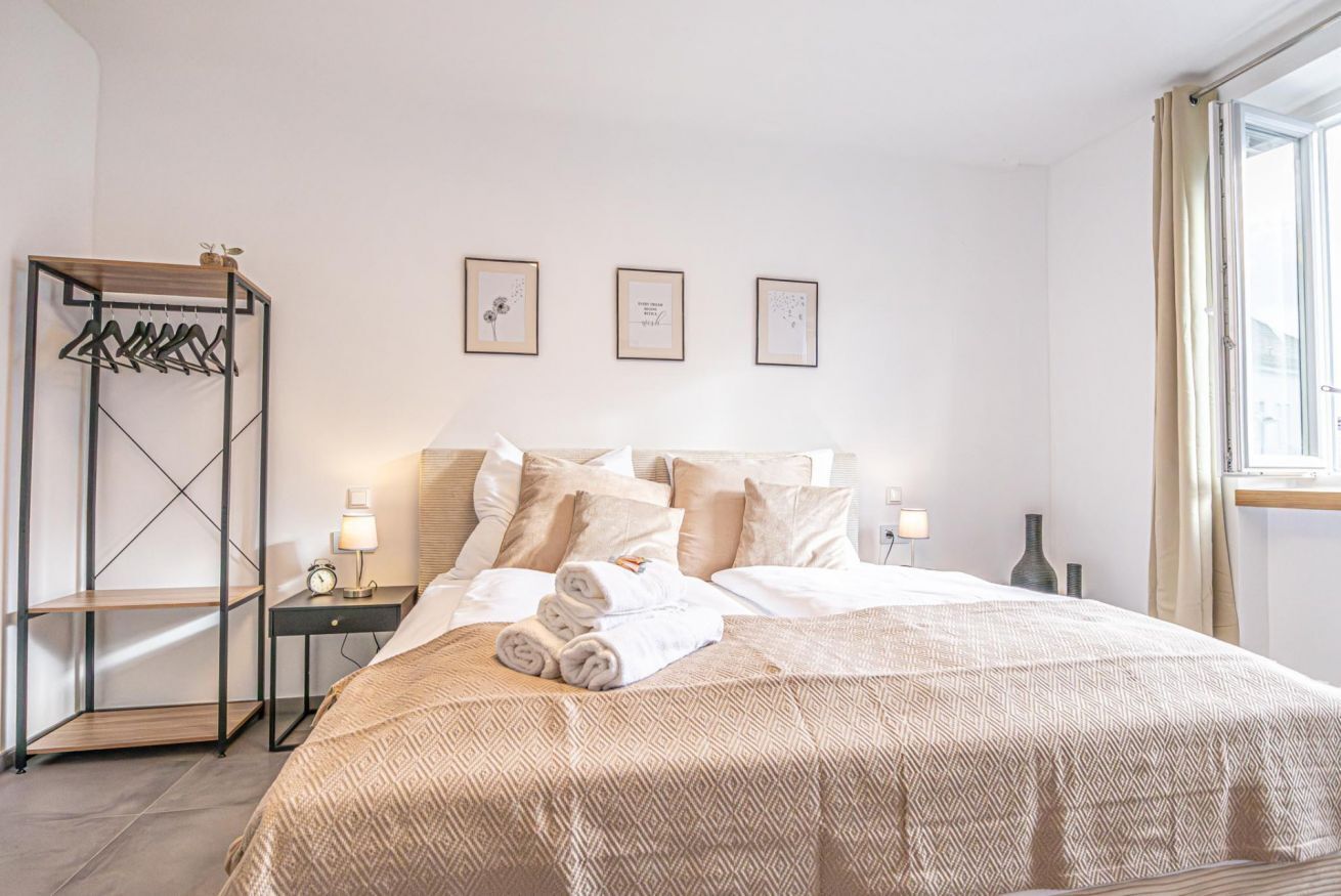 A neatly arranged bedroom with a bed, bedside tables, a hanging coat rack, and framed pictures on the wall.