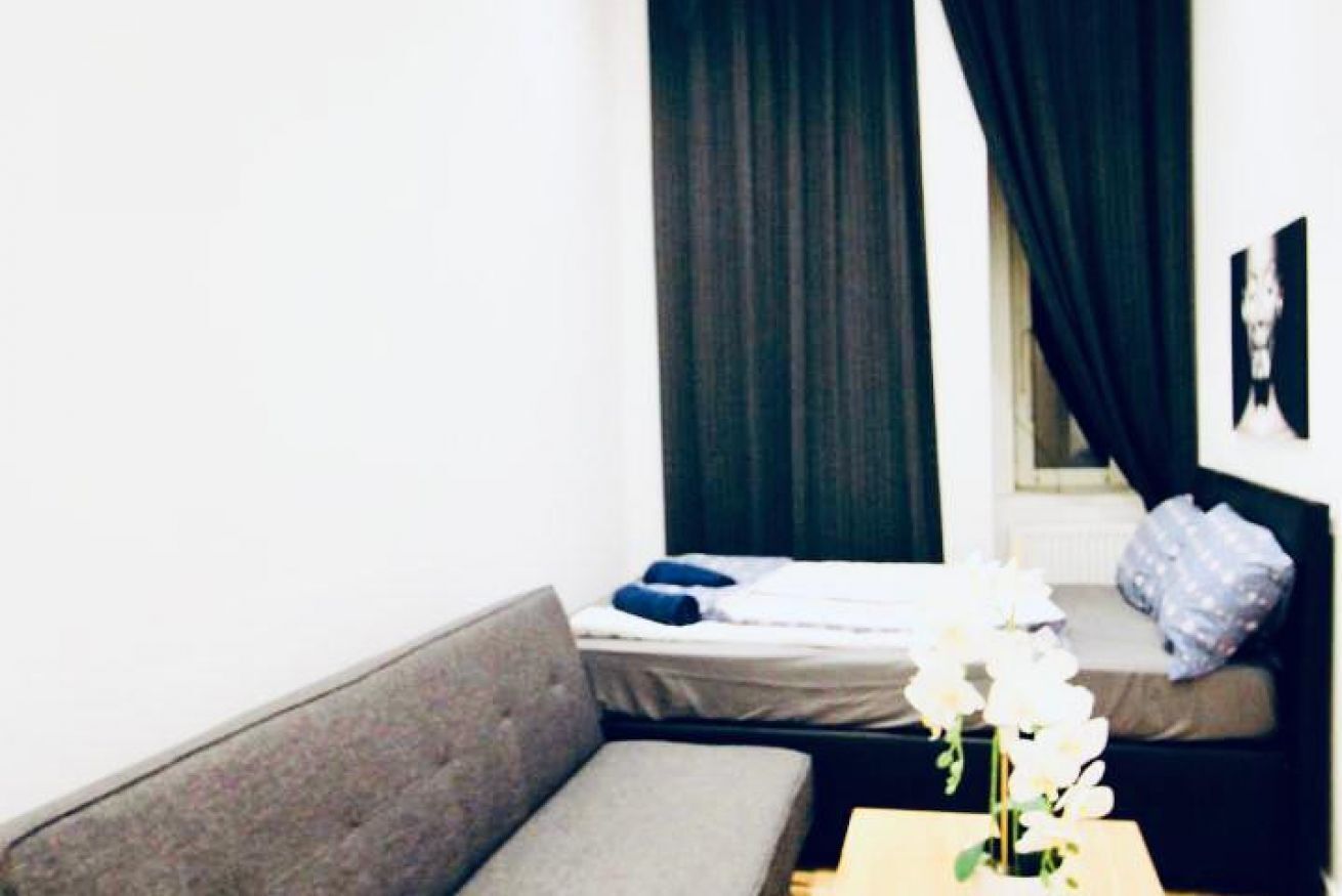 Una habitación con sofá, cama con almohada, una mesita con una flor y ventana con cortinas.
