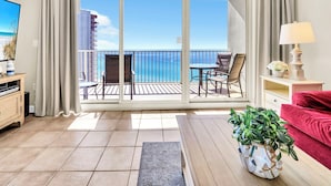 A balcony with a view of the ocean, a dining table with chairs, a red sofa, a TV, and a lamp. - 🌊Gulf and Pool Views-Pool-Fab Amenities-Shores of Panama 1711🌊 (Panama City Beach)