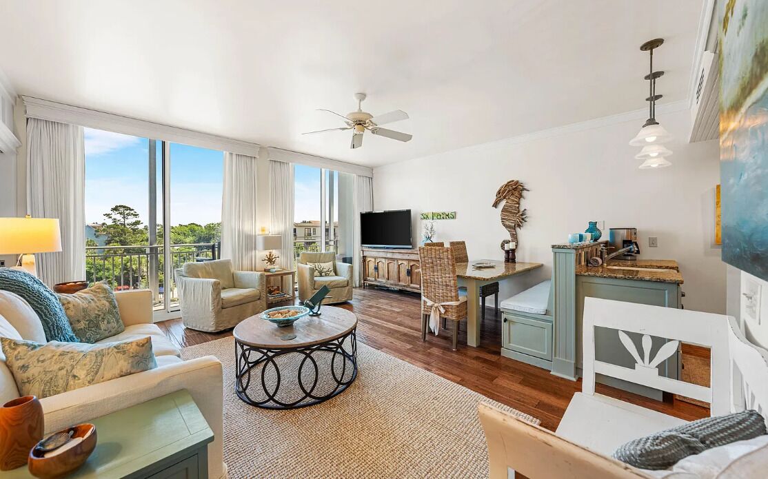 A spacious living room with a large window, a ceiling fan, a television, and a dining area with a table and chairs.