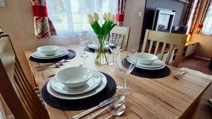 A dining area with a wooden table set for four, including plates, cutlery, and a vase of white flowers. - Caravan by the sea with direct access to the beach.  Pet friendly! (Barton on Sea)