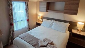 A hotel room with a large bed, two bedside tables, a window with curtains, and a wooden headboard. - Caravan by the sea with direct access to the beach.  Pet friendly! (Barton on Sea)