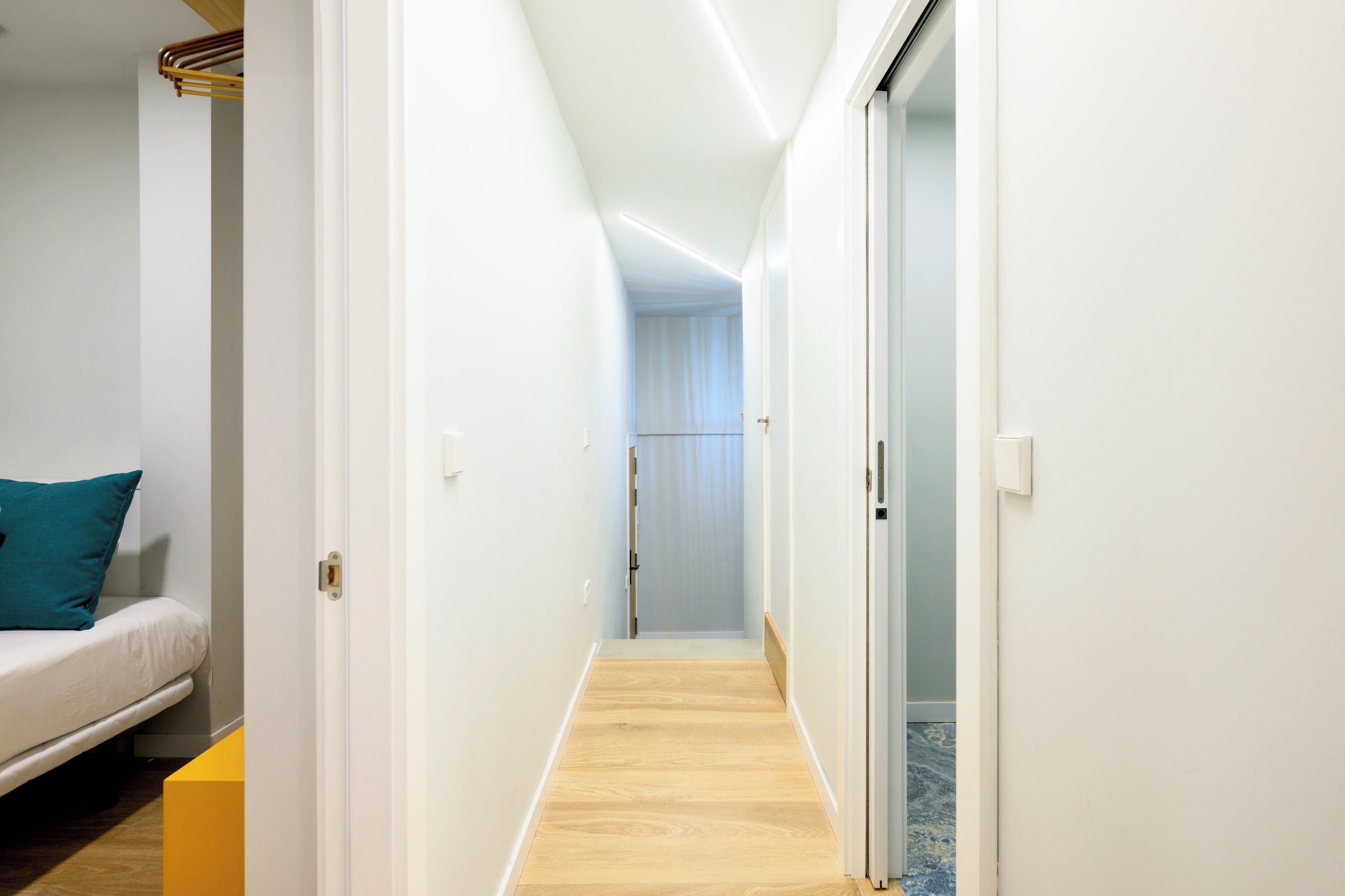 Een smalle gang met een houten vloer, witte muren en een deur die leidt naar een kamer met een bed en een blauw kussen.