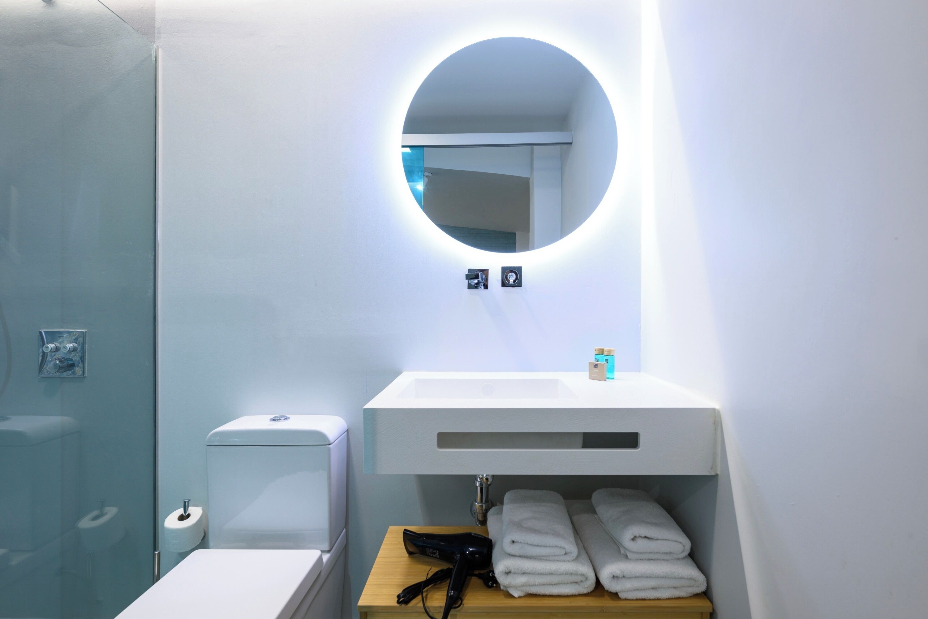 A modern bathroom with a round mirror, white sink, and neatly folded towels on a wooden shelf.