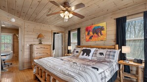 Une chambre dans une cabane en bois, avec un lit, un bureau, une chaise, un ventilateur de plafond et un tableau accroché au mur.
