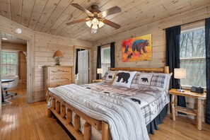 A wooden cabin bedroom with a bed, a desk, a chair, a ceiling fan, and a painting on the wall.