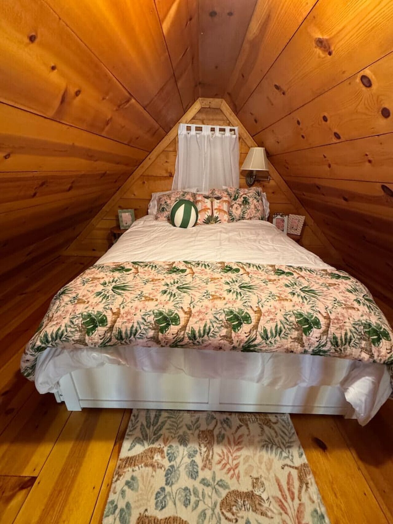Un dormitorio acogedor en el ático con techo inclinado de madera, una cama con ropa de cama floral, dosel y una mesita de noche pequeña.