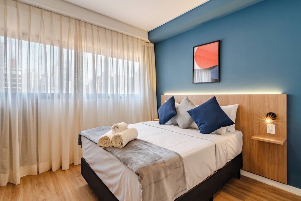 A modern bedroom with a bed, curtains, a wall-mounted light, and a city view.