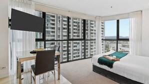 A modern hotel room with a large bed, a desk with a chair, a TV mounted on the wall, and a view of a cityscape through large windows. - Seabright Nook Resort Perks and Sky Views by the Sea (Surfers Paradise)
