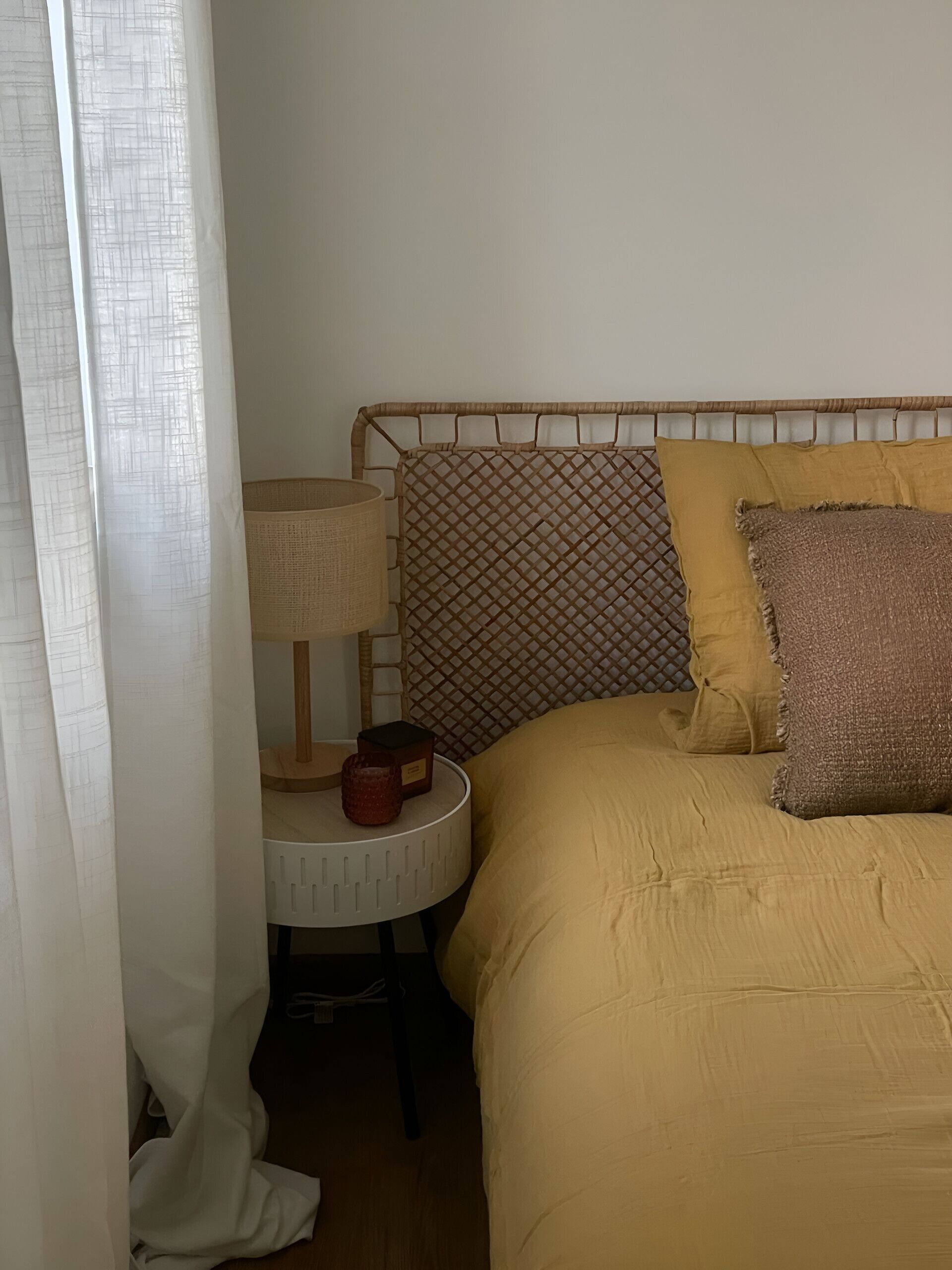A bed with a yellow duvet, a woven headboard, and two pillows. There is a bedside table with a lamp and a small box.