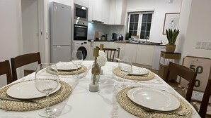 Zona pranzo con tavolo bianco apparecchiata per quattro persone, circondata da sedie. In secondo piano, si intravede una cucina con mobili ed elettrodomestici bianchi.