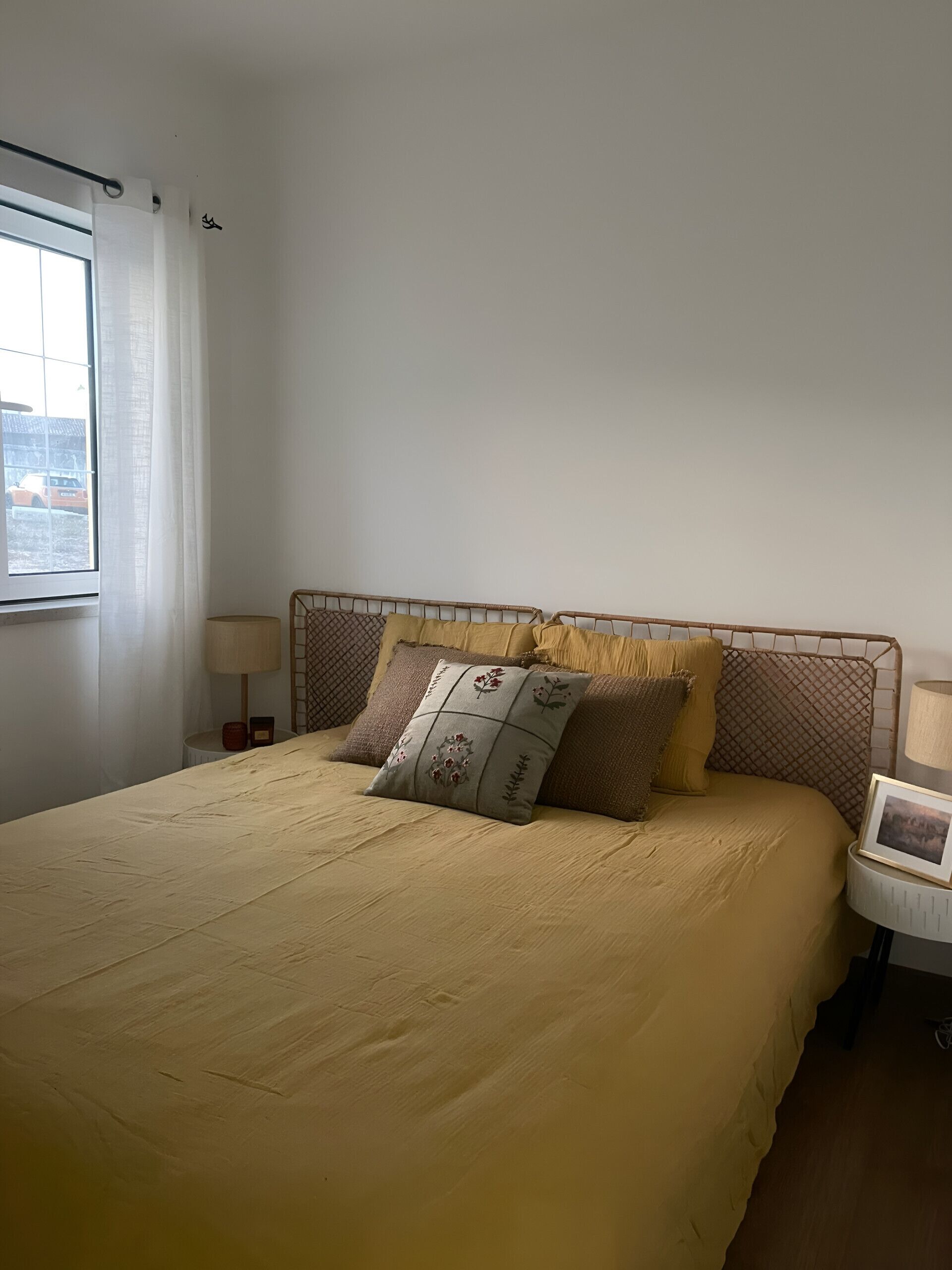 Un lit bien fait, recouvert d’une couvre-lit beige et agrémenté de plusieurs coussins, une table de chevet avec une lampe, et une fenêtre avec un rideau.