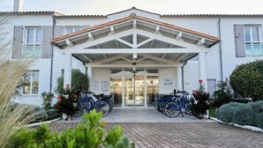 A building with a white facade, a tiled roof, and a covered entrance with glass doors. There are bicycles parked in front. - THALACAP Ile de Ré (Ars-en-Ré)
