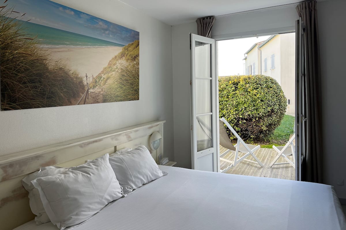 A bedroom with a bed, white pillows, a painting of a beach, and an open door leading to a balcony with a chair and a hedge.