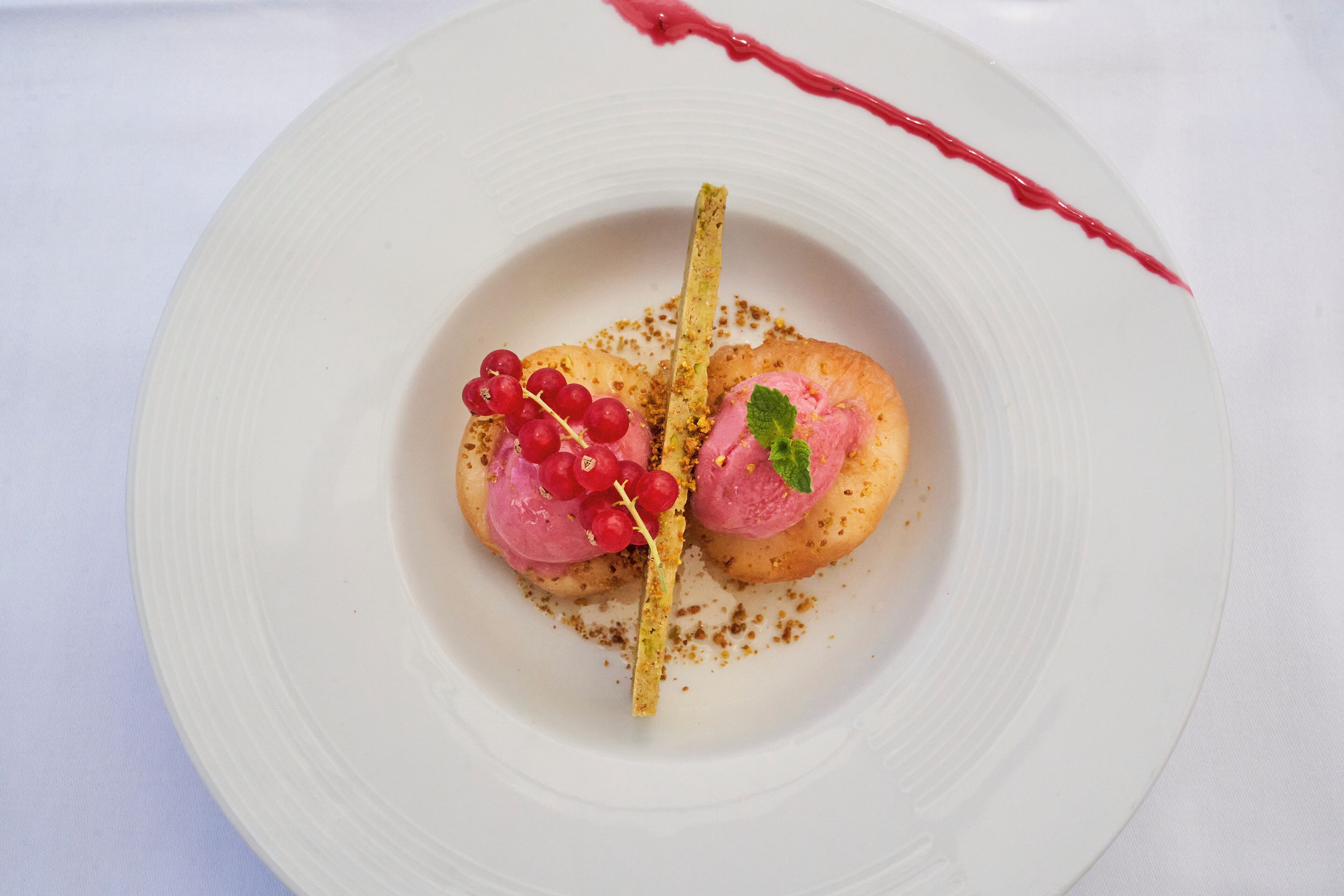 Une assiette blanche contenant un dessert composé de deux moitiés d’une pâtisserie, garnies de groseilles rouges et d’une feuille de menthe, accompagné d’un fin bâtonnet jaune.