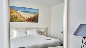 Une chambre d’hôtel avec un lit, un bureau doté d’une télécommande, une lampe et un tableau représentant une plage.