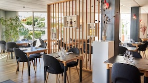 A modern restaurant with wooden tables and chairs, a decorative partition, and a view of greenery outside. - THALACAP Ile de Ré (Ars-en-Ré)