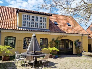 Ein gelbes Gebäude mit einem Ziegeldach und einem großen Fenster. Vor der Fassade befindet sich ein runder Tisch mit Stühlen und ein Sonnenschirm.