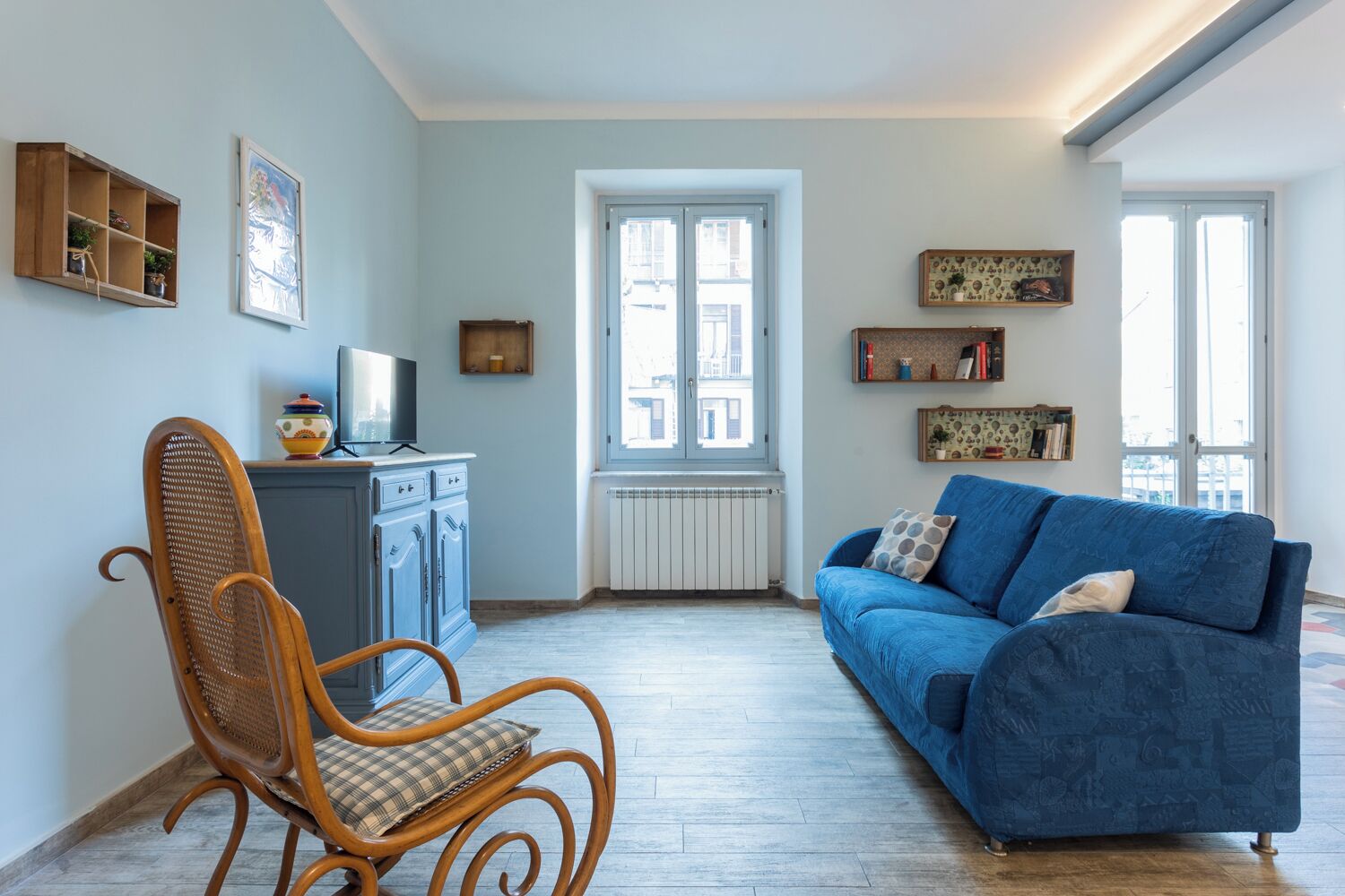 A living room with a blue sofa, a wicker chair, a cabinet with a TV, and a window with a view of buildings.