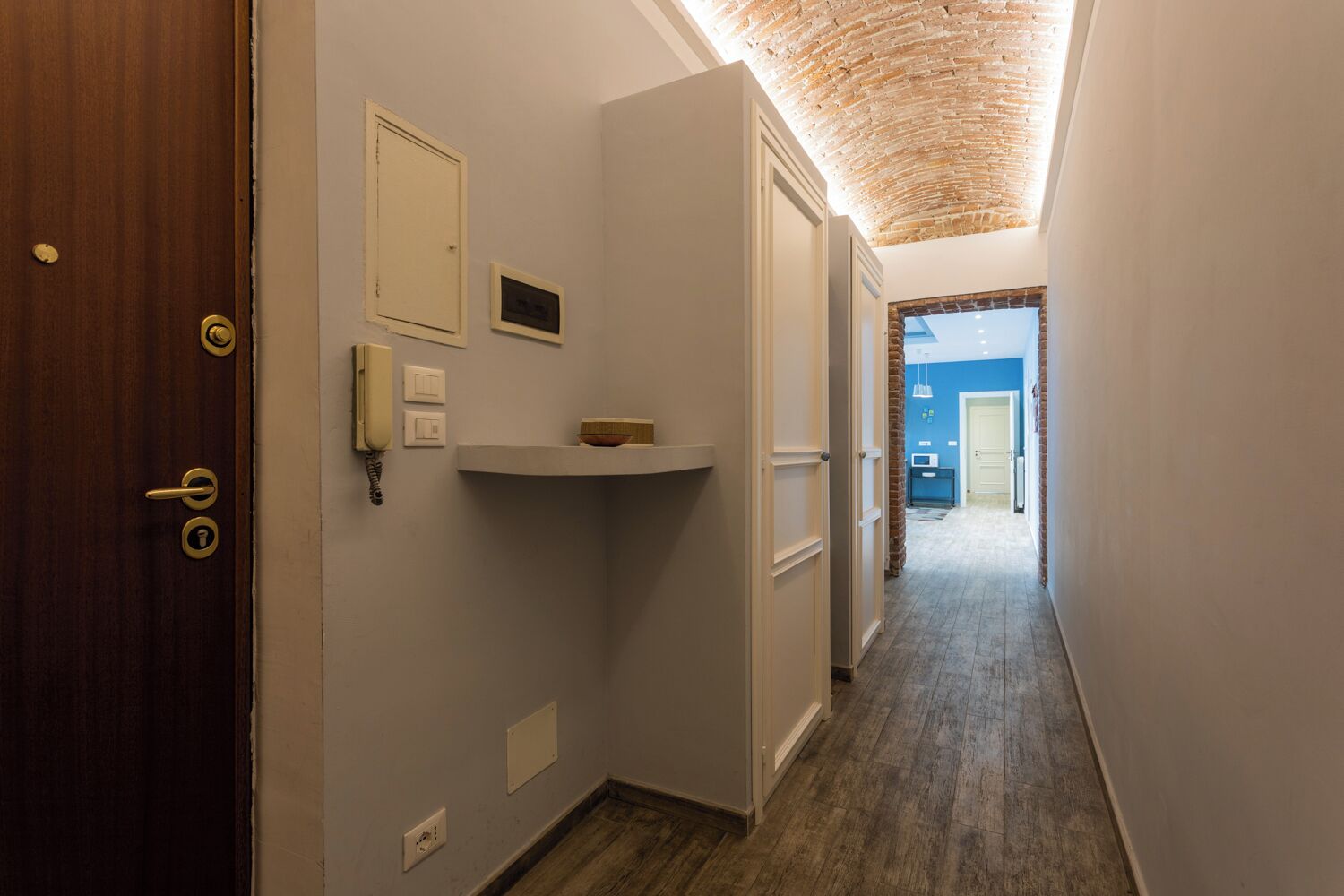 A narrow hallway with a wooden door on the left, a white shelf with a bowl, and a blue wall at the end.