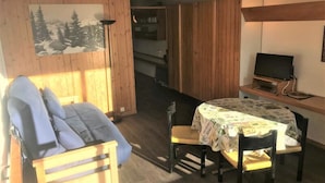 Een gezellige kamer met een houten vloer, een bank, een eettafel met stoelen, een televisie op een meubelstuk en een schilderij van een besneeerd landschap.