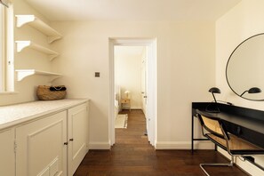A hallway with white walls, a wooden floor, and a door leading to another room. There is a small table with a lamp and a basket on the left side.
