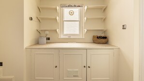 A compact room with a built-in kitchenette, a window with a clock, and a small shelf unit with baskets. - Notting Hill Ground-Floor Retreat (London)