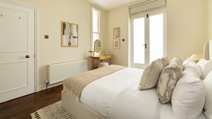 A bedroom with a large bed, a dresser, a mirror, and two windows. - Notting Hill Ground-Floor Retreat (London)