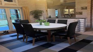 A dining area with a round table, black chairs, and a green bowl centerpiece. There is a large window with curtains, a potted plant, and a kitchen area visible through an open door. - 4 Star Holiday Home in Ronne (Ronne)