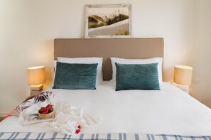 A neatly made bed with white and teal pillows, a framed picture above, and a bedside table with a lamp.