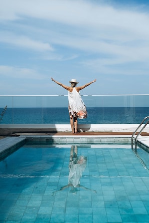 A person standing on a pool deck with arms outstretched, overlooking a pool and the ocean.
