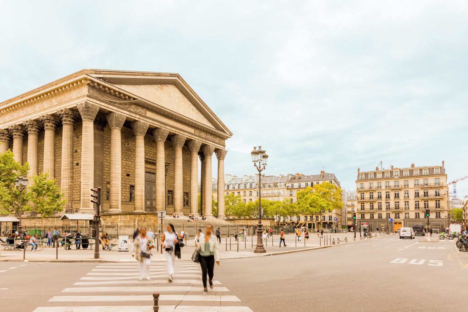 A large classical building with columns, a crosswalk, and a busy street with pedestrians and vehicles.