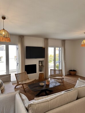 A living room with a sofa, two wicker chairs, a coffee table, a TV mounted on the wall, and a large window with curtains.