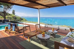 Eine Holzterrasse mit Sitzgelegenheiten im Freien, einem Pool und Blick aufs Meer.