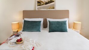 Ein ordentlich bezogenes Bett mit weißen und türkisblauen Kissen, ein gerahmtes Bild darüber und ein Nachttischchen mit einer Lampe.
