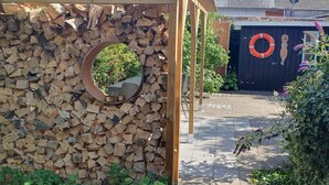 Una pergola de fusta amb un mirall circular, una pila de llenya, i un jardí ple de plantes i flors verdes.