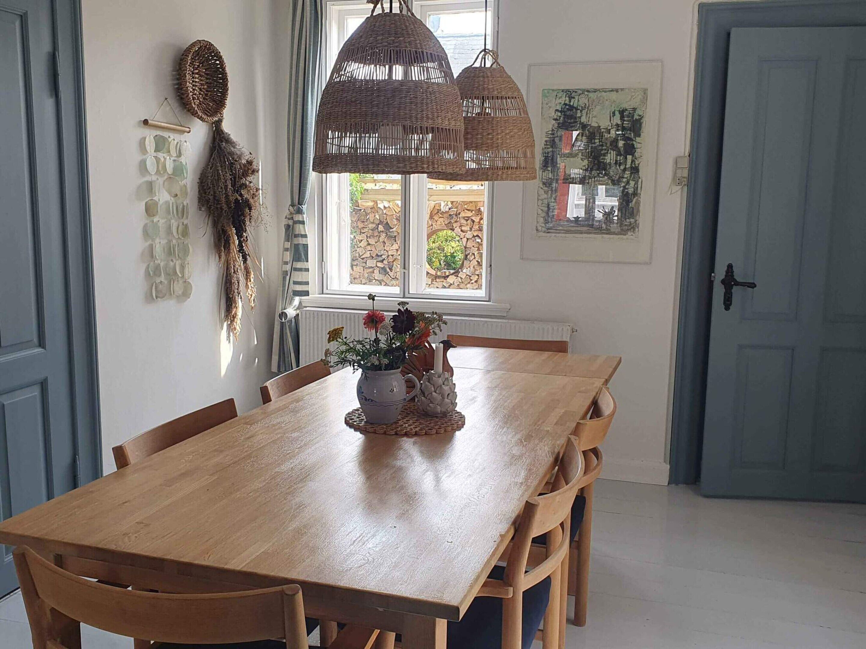 Une table à manger en bois, accompagnée de quatre chaises, une suspension en fibres tressées et un vase rempli de fleurs.