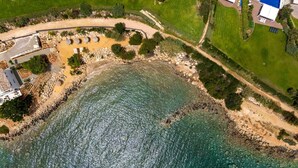 Vue aérienne d’une propriété côtière avec une plage de sable, une côte rocheuse et une mer d’un bleu limpide. On aperçoit plusieurs structures circulaires près du bord de l’eau, probablement destinées à des activités de loisirs.