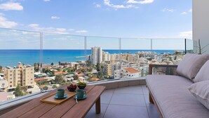 A balcony with a view of the sea and cityscape, a wooden table with a tray, and a sofa. - Suite 1003 at Althea Residence (Larnaca)