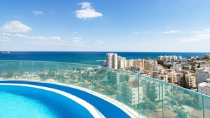 A high-rise building with a glass balcony overlooking a swimming pool and a coastal cityscape. - Suite 1003 at Althea Residence (Larnaca)