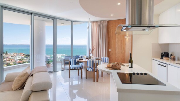 A modern kitchen with a central island, a dining area with blue-striped chairs, and a large sliding glass door offering a view of the sea. - Suite 1003 at Althea Residence (Larnaca)