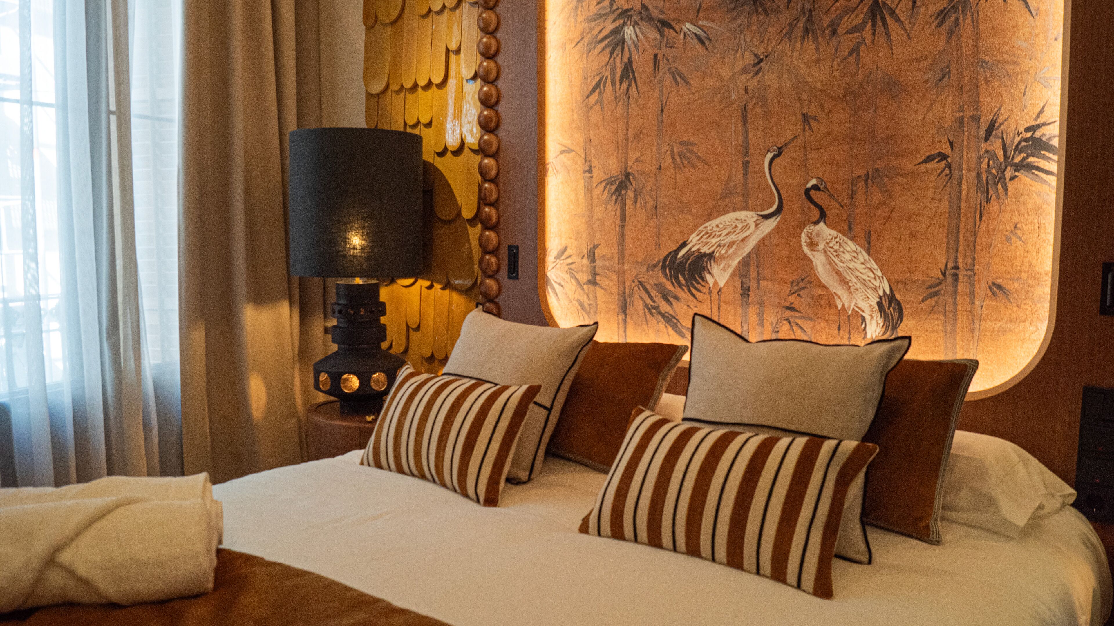 Habitación de hotel con cama, almohadas a rayas, mesita de noche con lámpara y un mural de bosque de bambú en la pared.