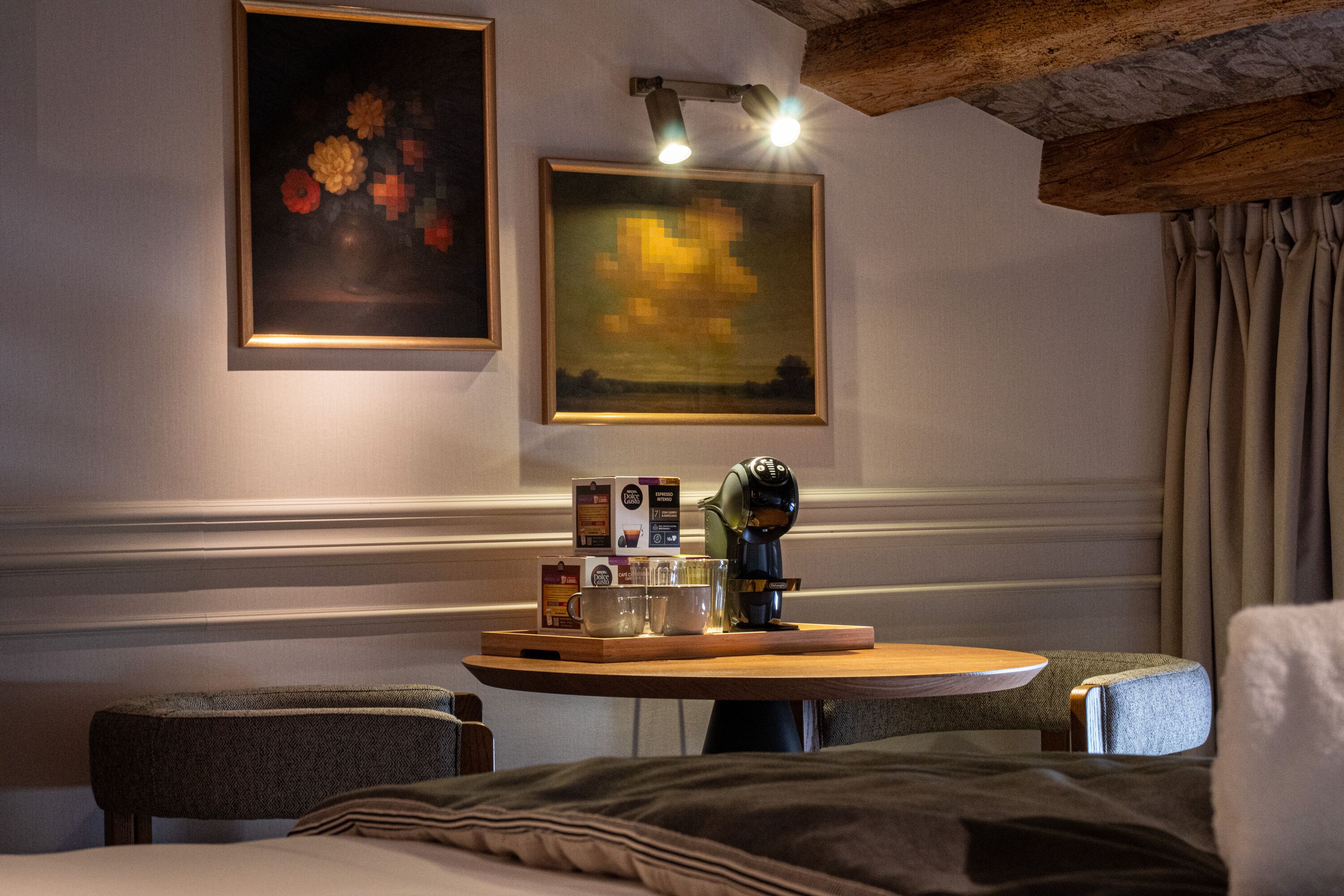 Une chambre d’hôtel confortable avec un lit, une petite table ronde sur laquelle se trouvent une cafetière, des tasses et une boîte de thé. Deux tableaux encadrés ornent le mur, et une poutre en bois traverse le plafond.