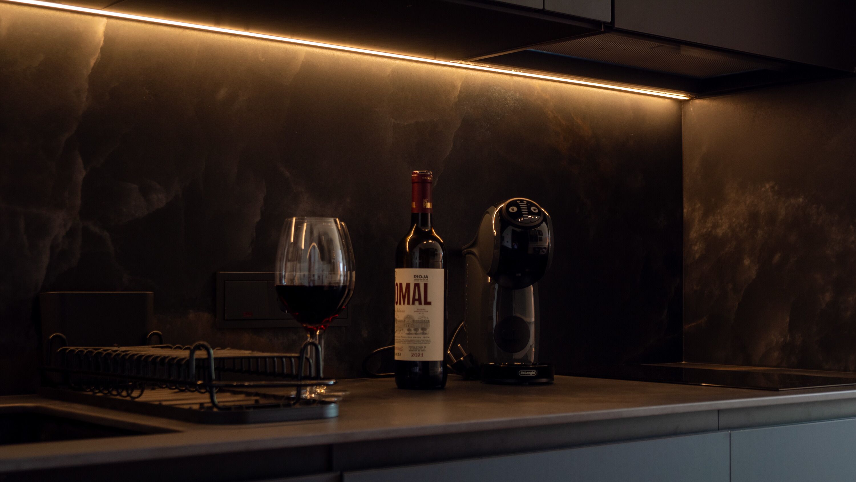 A kitchen counter with a wine bottle, a glass of red wine, and a coffee machine.